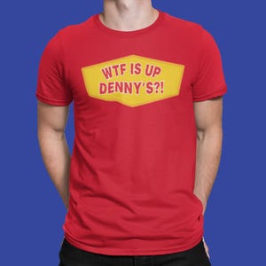 May include: Red t-shirt with the text "WTF IS UP DENNY'S?!" in a yellow and red design. The text is inside a yellow, diamond-shaped graphic. The shirt is short-sleeved.
