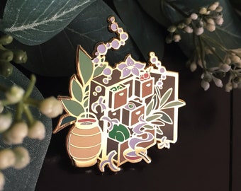 May include: Gold-tone enamel pin featuring a whimsical illustration of a small house with a chimney, surrounded by flowers and vines. The house has a series of drawers with different colored accents.