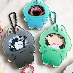 May include: A selection of keychains with cartoon character designs. The keychains are shaped like furry monsters in blue, grey, and green. Each has a ball chain and a character image within the monster suit.