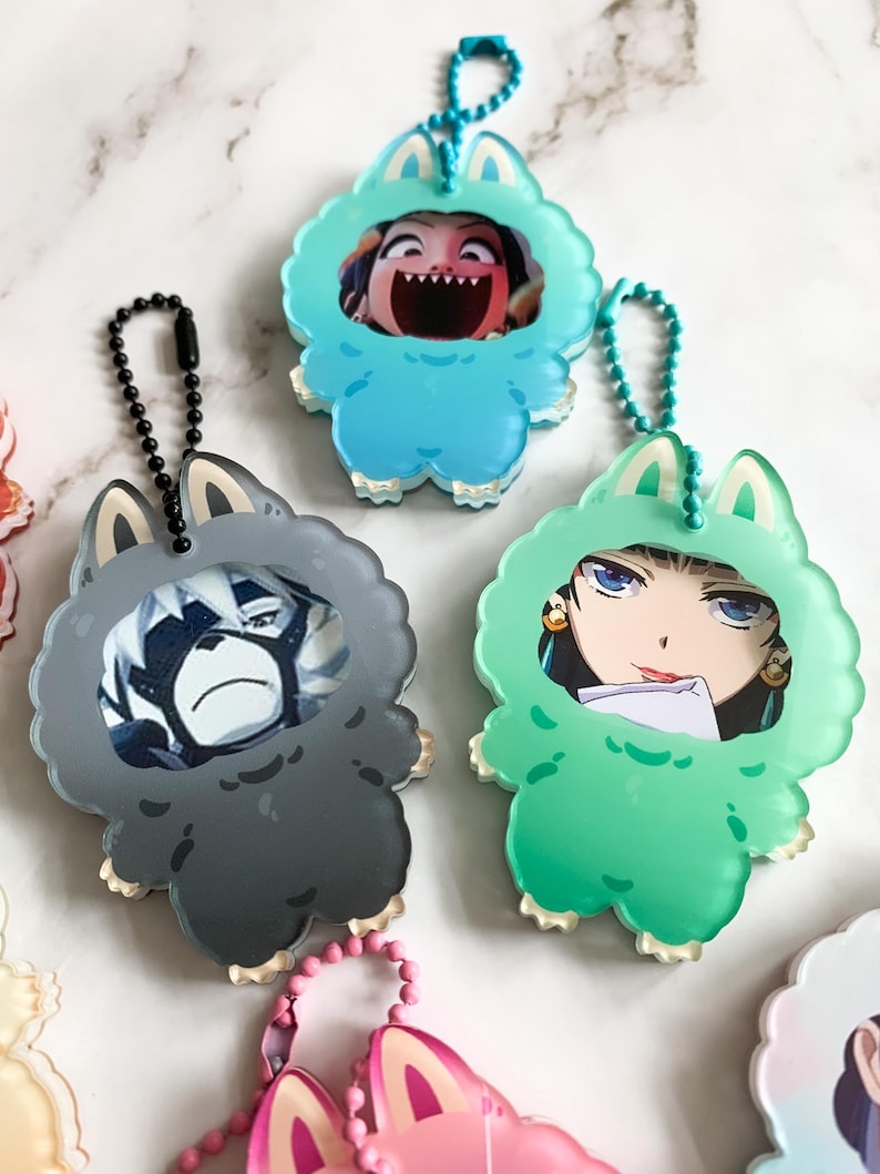 May include: A selection of keychains with cartoon character designs. The keychains are shaped like furry monsters in blue, grey, and green. Each has a ball chain and a character image within the monster suit.