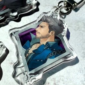 May include: A clear acrylic keychain featuring a cartoon illustration of a man with grey hair and a beard. He is wearing a blue jacket with a white shirt underneath. The image is set against a purple background and framed with a silver border.