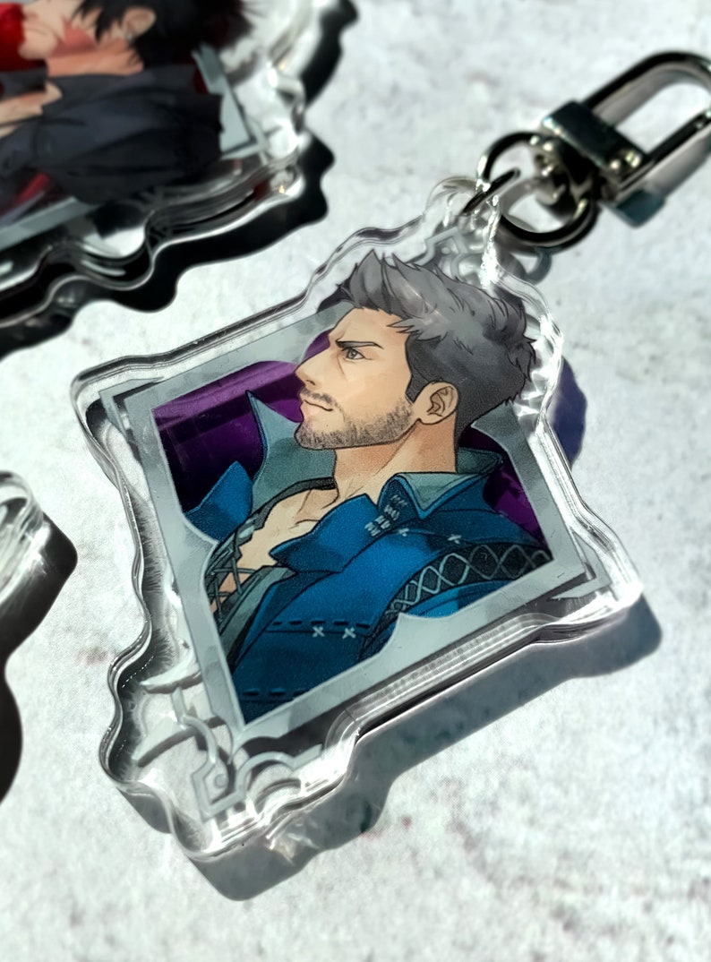 May include: A clear acrylic keychain featuring a cartoon illustration of a man with grey hair and a beard. He is wearing a blue jacket with a white shirt underneath. The image is set against a purple background and framed with a silver border.