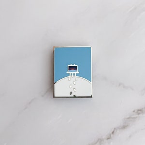 May include: A silver enamel pin with a white background and a blue sky. The pin features a white easel with a colourful sunset painting on it. There are white dots scattered on the ground below the easel.
