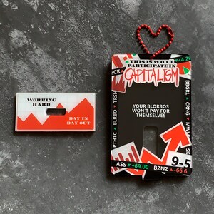 May include: A black plastic card with red and white graphics. The card has the text "This is why I participate in Capitalism" and "Your Blorbos won't pay for themselves". The card also has a red arrow pointing up with the text "9-5" and "ASS +69.00 BZNZ -66.6".