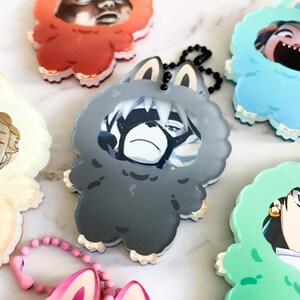 May include: A selection of cartoon keychains in various colours. Each keychain has a character design within a fluffy animal-shaped frame. The keychains are in shades of red, grey, blue, green, pink, and yellow, with black ball chain attachments.