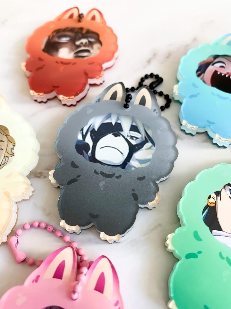 May include: A selection of cartoon keychains in various colours. Each keychain has a character design within a fluffy animal-shaped frame. The keychains are in shades of red, grey, blue, green, pink, and yellow, with black ball chain attachments.