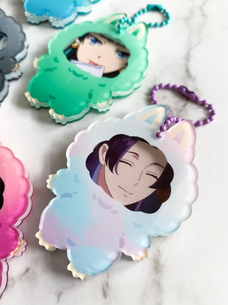 May include: A selection of anime-style character keyrings. Each keyring features a character portrait within a fluffy, animal-shaped frame. The frames are in various pastel colours, including green, blue, pink, and white, with a matching chain.