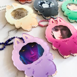 May include: A selection of colourful acrylic charms, each depicting a character portrait within a fluffy animal-shaped frame. The charms, in various colours, have a keychain attachment and are displayed on a white surface.