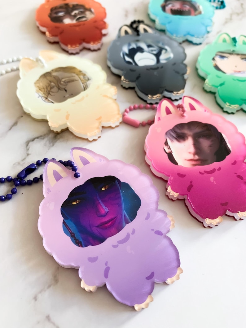 May include: A selection of colourful acrylic charms, each depicting a character portrait within a fluffy animal-shaped frame. The charms, in various colours, have a keychain attachment and are displayed on a white surface.