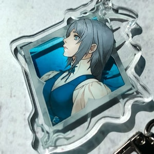May include: A clear acrylic keychain featuring a portrait of a woman with silver hair and a blue dress. The image is framed in a silver frame with a decorative border.