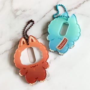 May include: Two acrylic keychains. One is orange with a fox-like design, the other is blue and teal with a cloud-like design. Both have a rectangular opening and a chain for attachment. The blue keychain has the word "BOBMART" printed on it.