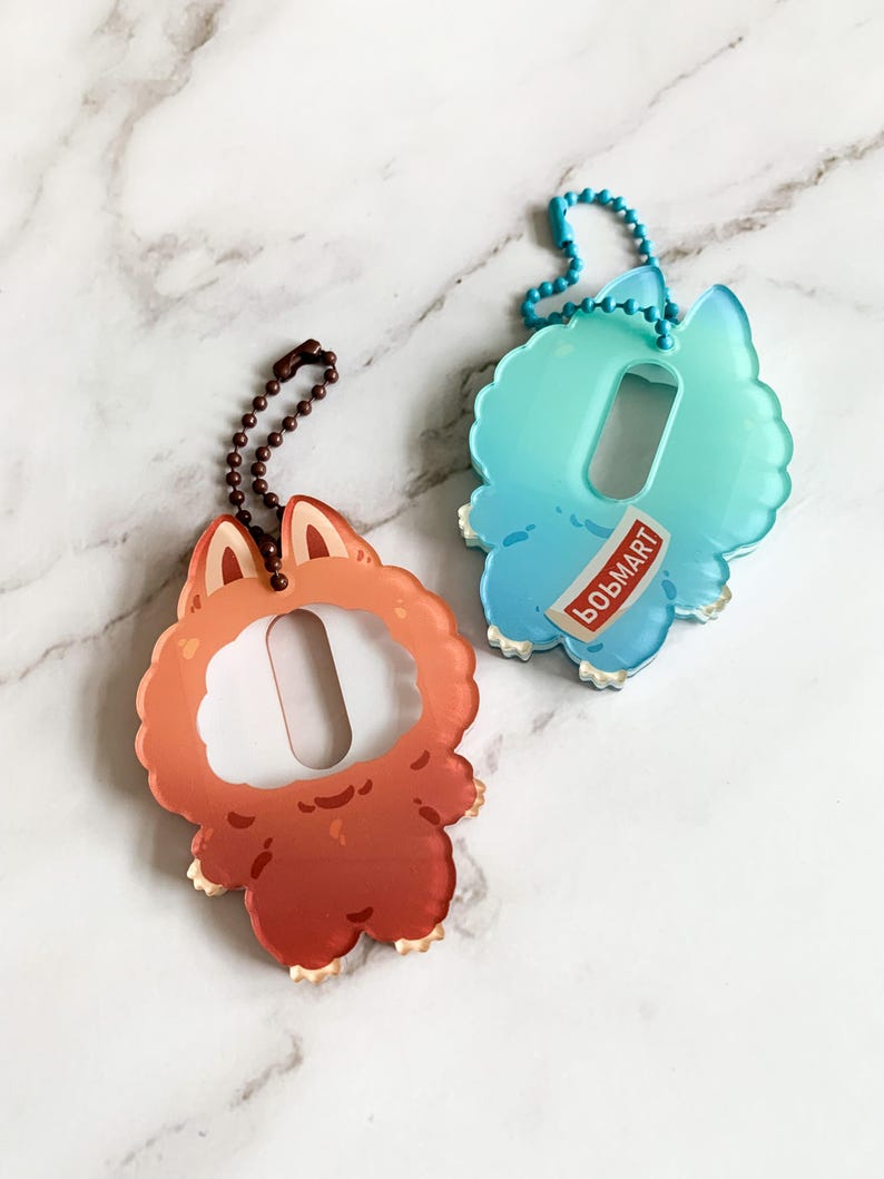 May include: Two acrylic keychains. One is orange with a fox-like design, the other is blue and teal with a cloud-like design. Both have a rectangular opening and a chain for attachment. The blue keychain has the word "BOBMART" printed on it.