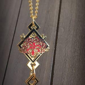 May include: A gold necklace with a black and red enamel pendant featuring a stylized flower design. The pendant is shaped like a diamond and has a geometric pattern.