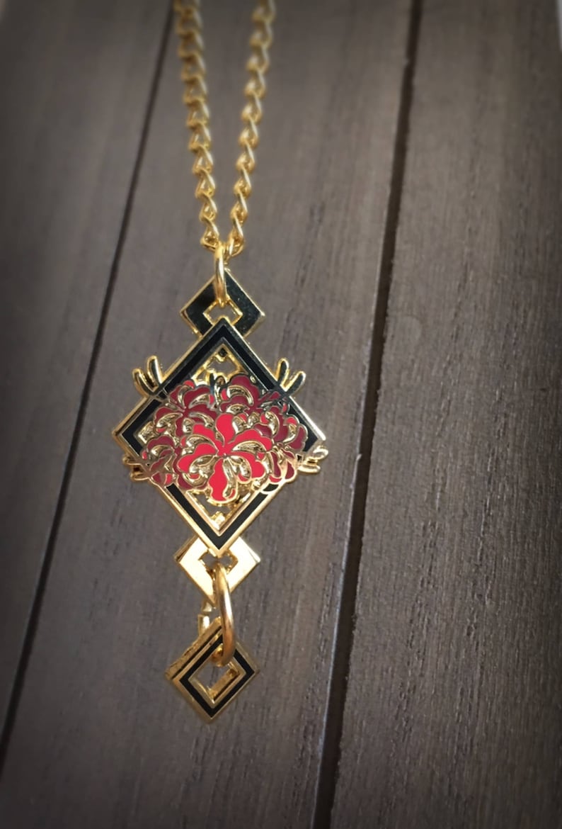 May include: A gold necklace with a black and red enamel pendant featuring a stylized flower design. The pendant is shaped like a diamond and has a geometric pattern.