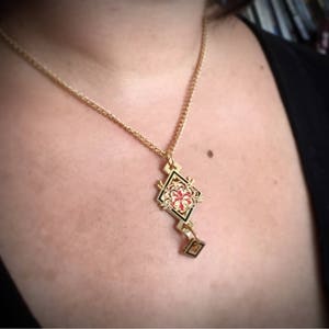 May include: A gold necklace with a geometric pendant featuring a red and black floral design.