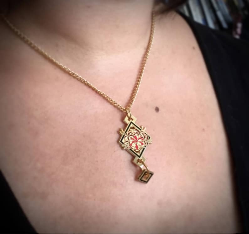 May include: A gold necklace with a geometric pendant featuring a red and black floral design.