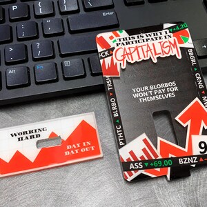 May include: Two red and white plastic cards with a graphic of a rising stock market. The text on the cards reads "Working Hard Day In Day Out" and "This is why I participate in Capitalism".
