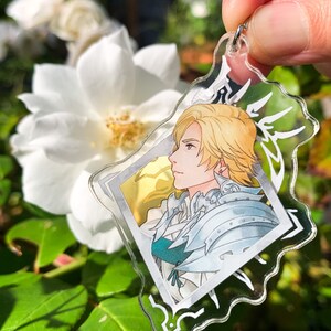 May include: A clear acrylic keychain featuring a character with blond hair and a white and blue outfit. The character is depicted in a profile view, looking to the right. The keychain has a silver metal ring.