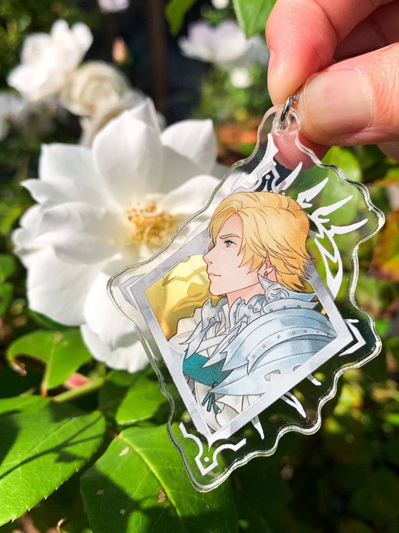 May include: A clear acrylic keychain featuring a character with blond hair and a white and blue outfit. The character is depicted in a profile view, looking to the right. The keychain has a silver metal ring.