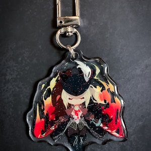 May include: A clear acrylic keychain featuring a cartoon character with blonde hair, a black mask, and a white feather. The character is surrounded by red and orange flames and glitter.