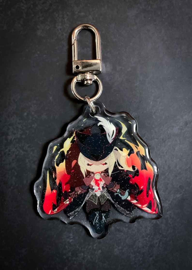 May include: A clear acrylic keychain featuring a cartoon character with blonde hair, a black mask, and a white feather. The character is surrounded by red and orange flames and glitter.