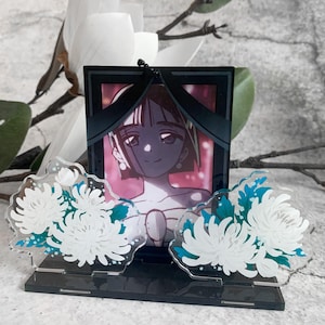 May include: A black acrylic memorial frame with a cartoon illustration of a person, adorned with black ribbons. Two clear acrylic flower ornaments with white and teal accents flank the frame, creating a poignant display.