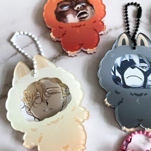 May include: A selection of anime-style character keychains. Each keychain has a character portrait inside a fluffy animal-shaped frame. The keychains are in various colours, including orange, cream, and grey, with matching chain attachments.