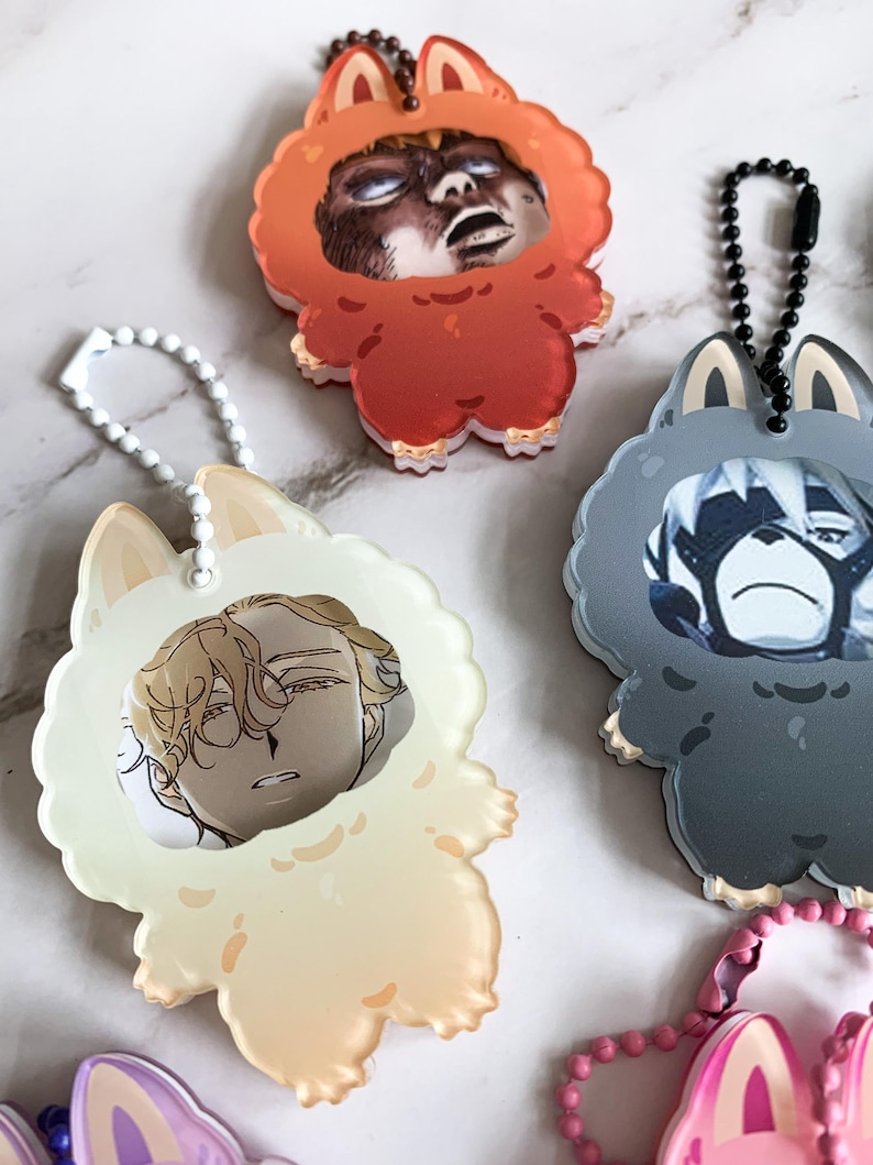 May include: A selection of anime-style character keychains. Each keychain has a character portrait inside a fluffy animal-shaped frame. The keychains are in various colours, including orange, cream, and grey, with matching chain attachments.