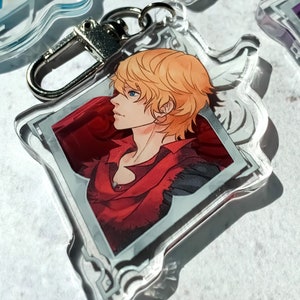 May include: A clear acrylic keychain featuring a character with blond hair and a red shirt. The character is depicted in profile, looking to the left. The keychain has a silver metal ring.