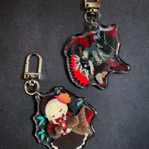 May include: Two character charms with glitter accents. One charm features a black and red character with a saw blade. The other charm features a female character with a red and brown dress and a black hat.