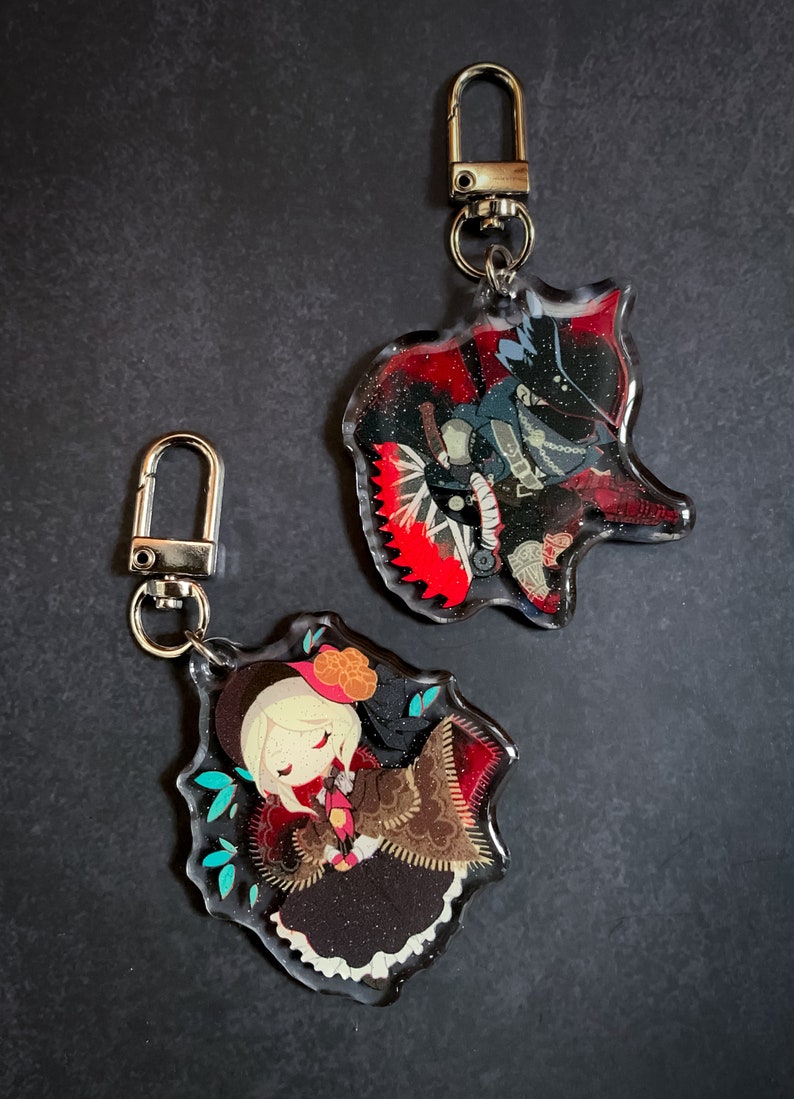May include: Two character charms with glitter accents. One charm features a black and red character with a saw blade. The other charm features a female character with a red and brown dress and a black hat.
