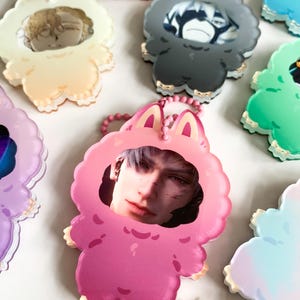 May include: A selection of acrylic keyrings in various colours. Each keyring features a portrait of a person within a cartoon animal-shaped frame. The frames are in shades like pink, grey, and green, with gold trim and a matching chain.