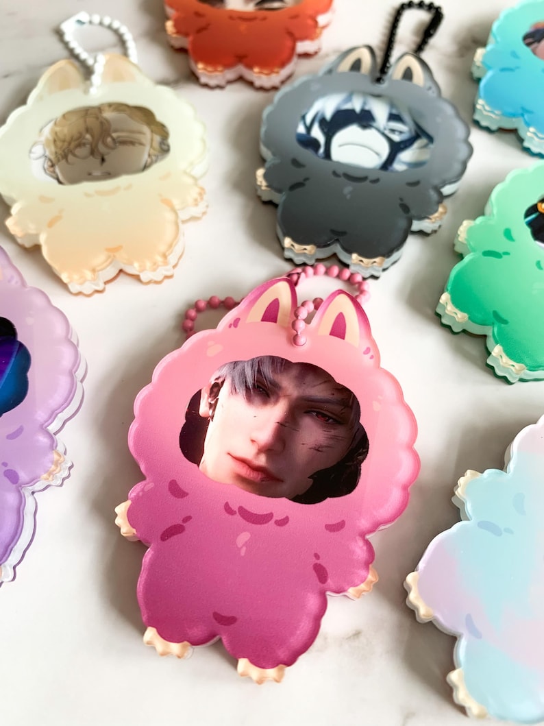May include: A selection of acrylic keyrings in various colours. Each keyring features a portrait of a person within a cartoon animal-shaped frame. The frames are in shades like pink, grey, and green, with gold trim and a matching chain.