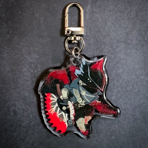 May include: A clear acrylic keychain featuring a cartoon character with a black and red costume, holding a weapon with a red blade. The character has a black mask and a white hat.