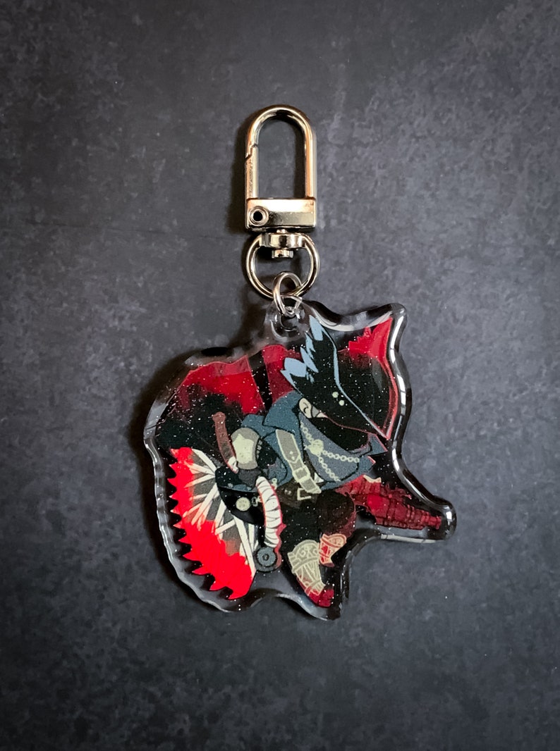 May include: A clear acrylic keychain featuring a cartoon character with a black and red costume, holding a weapon with a red blade. The character has a black mask and a white hat.