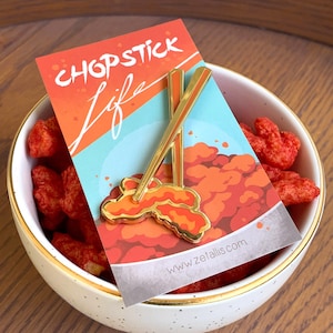 Può includere: Spilla in smalto dorata con un paio di bacchette con un design a forma di nuvola rosso e arancione. La spilla è su una carta con il testo "Chopstick Life" e il sito web "www.zetallis.com".