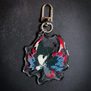 May include: A black and blue character with a white mask and red wings, printed on a clear acrylic keychain with a silver metal clasp.