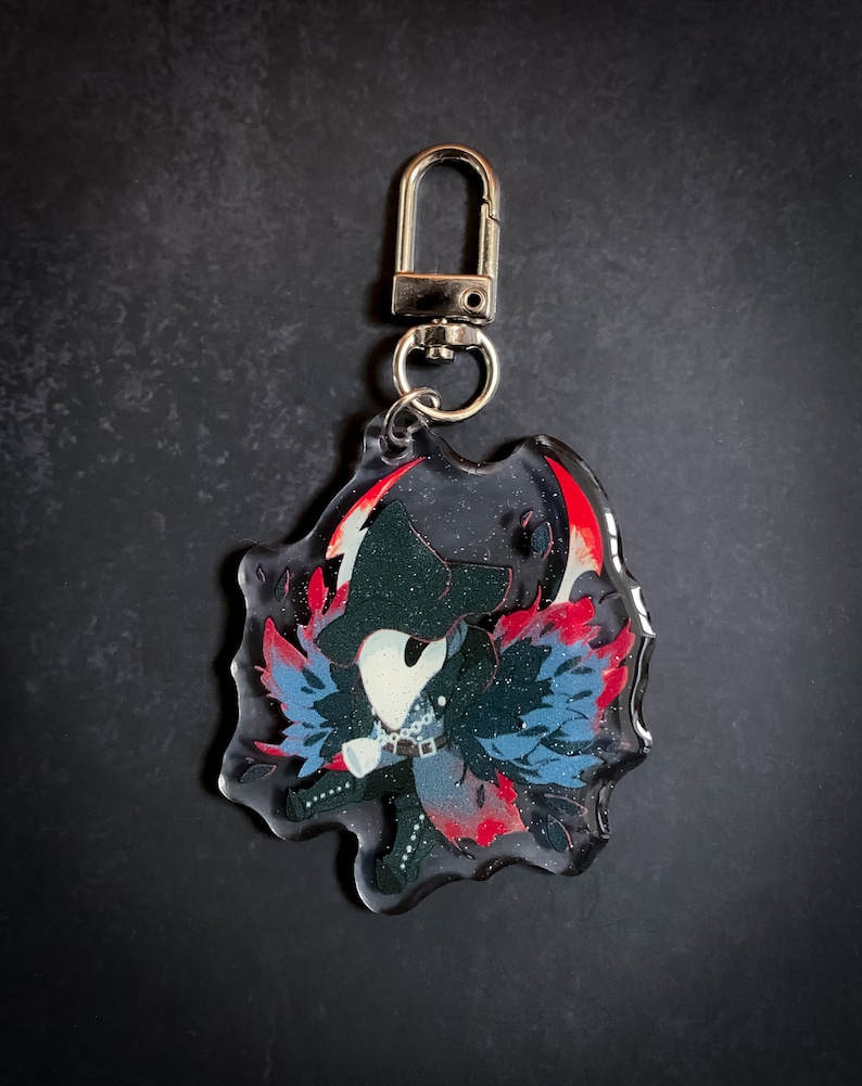 May include: A black and blue character with a white mask and red wings, printed on a clear acrylic keychain with a silver metal clasp.