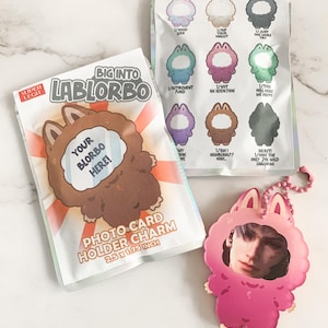 May include: A selection of Lablorbo-themed items: a photo card holder charm, a keychain, and a sticker sheet. The photo card holder measures 6.35 cm x 4.45 cm. The designs feature cartoon characters and text.