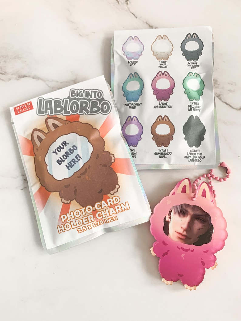 May include: A selection of Lablorbo-themed items: a photo card holder charm, a keychain, and a sticker sheet. The photo card holder measures 6.35 cm x 4.45 cm. The designs feature cartoon characters and text.