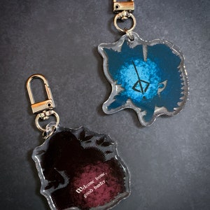May include: Two clear acrylic keychains with different designs. One keychain features a blue and white design with a symbol in the center. The other keychain features a black and red design with the text "Welcome home, good hunter".
