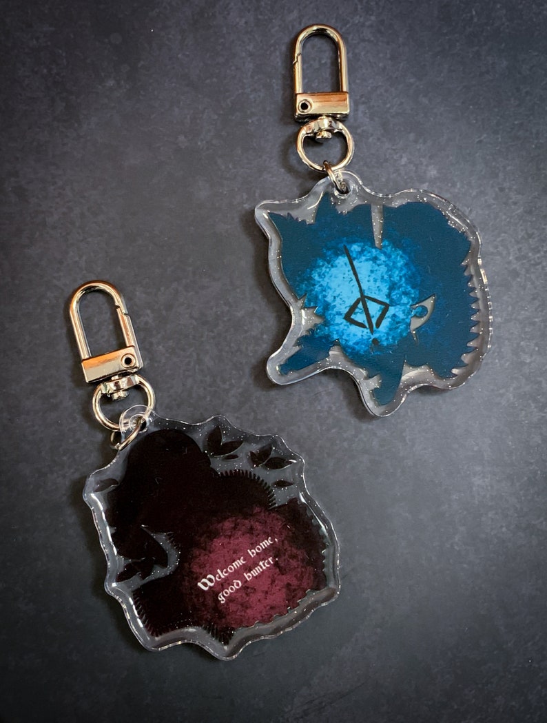 May include: Two clear acrylic keychains with different designs. One keychain features a blue and white design with a symbol in the center. The other keychain features a black and red design with the text "Welcome home, good hunter".