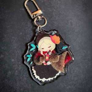 May include: A clear acrylic keychain featuring a cartoon character with blonde hair, wearing a brown and red dress with a black hat and a red bow. The character is surrounded by blue and green leaves.