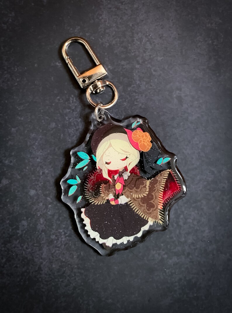 May include: A clear acrylic keychain featuring a cartoon character with blonde hair, wearing a brown and red dress with a black hat and a red bow. The character is surrounded by blue and green leaves.
