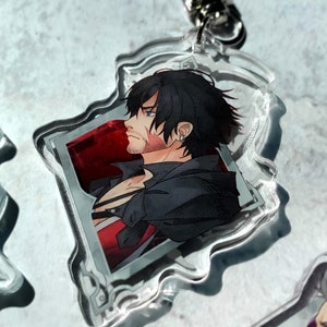 May include: A clear acrylic keychain featuring a character with black hair and a red and black outfit. The character is looking to the side and has a scar on their face. The keychain is shaped like a jagged rectangle with a silver metal ring.