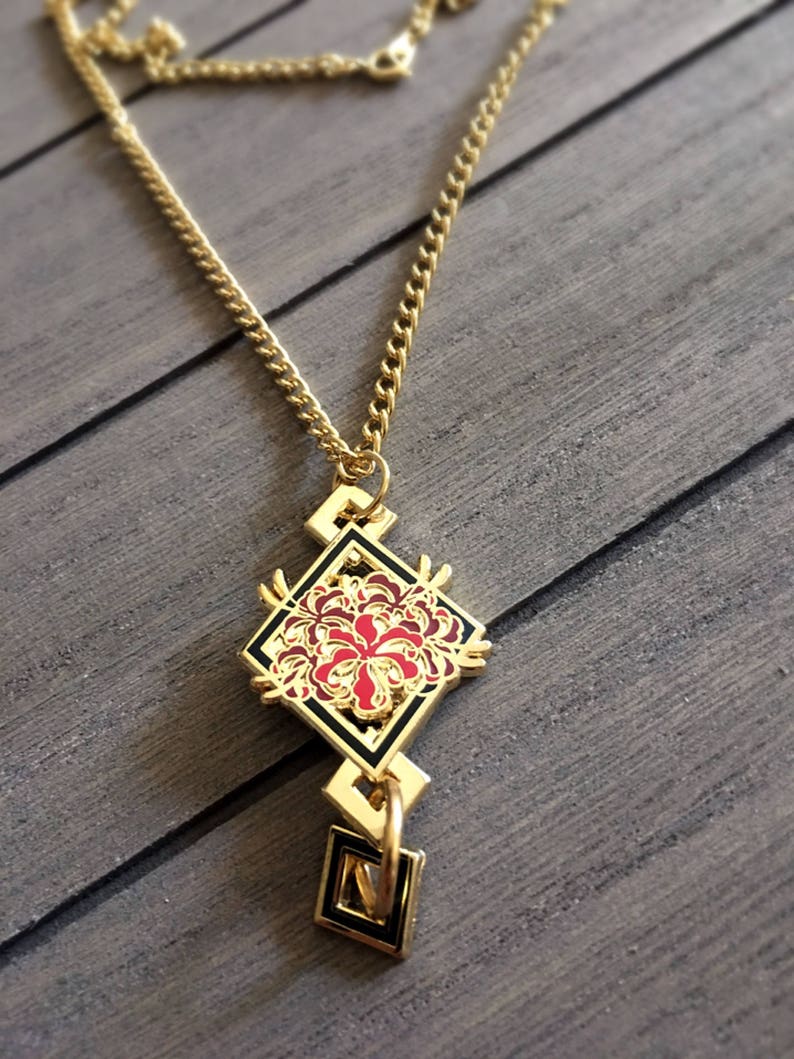 May include: A gold chain necklace with a pendant featuring a red and black stylized flower design. The pendant is in the shape of a diamond with a smaller diamond hanging below it.