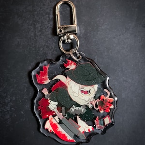 May include: A clear acrylic keychain featuring a cartoon character with a black hat, white fur, and red accents. The character is holding a red and black weapon and a small red box.