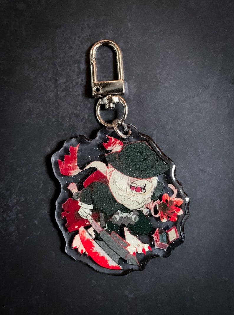 May include: A clear acrylic keychain featuring a cartoon character with a black hat, white fur, and red accents. The character is holding a red and black weapon and a small red box.