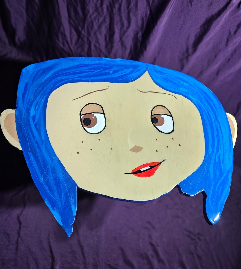 May include: A hand-painted wooden cutout of a girl with blue hair and a light brown complexion. The girl has brown eyes, a red mouth, and freckles.