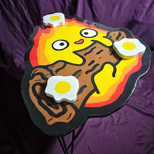 May include: A black and yellow painted wooden table shaped like a cartoon fire with a smiling face and three white eggs on top. The table is on a black metal stand with three legs.